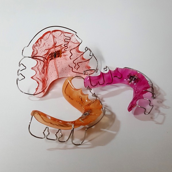 Drei herausnehmbare kieferorthopädische Geräte mit rosa, pinkem und orangefarbenem Kunststoffkörper und gebogenen Drahtklammern.