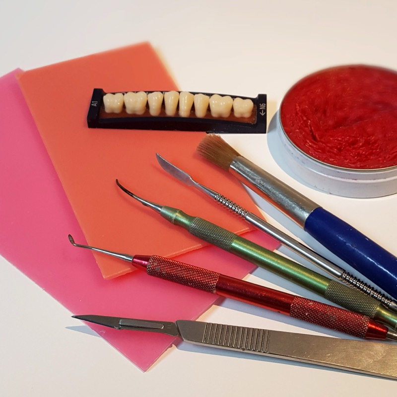 Arbeitsplatz im Labor: rosa Modellierwachs, eine Zahngarnitur der Farbe A1, Modellierinstrumente, Pinsel und ein Skalpell.