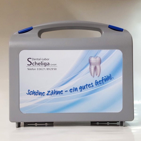 Grauer Transportkoffer des Labors mit blauem Aufkleber, Firmenlogo, Telefonnummer und dem Schriftzug „Schöne Zähne – ein gutes Gefühl.“